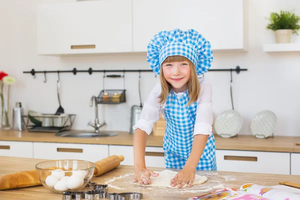 Pretty little girl in a cook clothes sculpts a circle of dough on a table and smiles