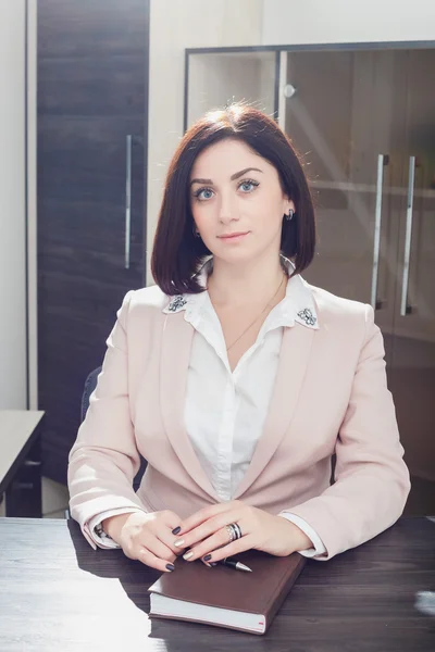 Attractive dark-haired woman dressed in a beige suit idit at  table in an office with  notebook.