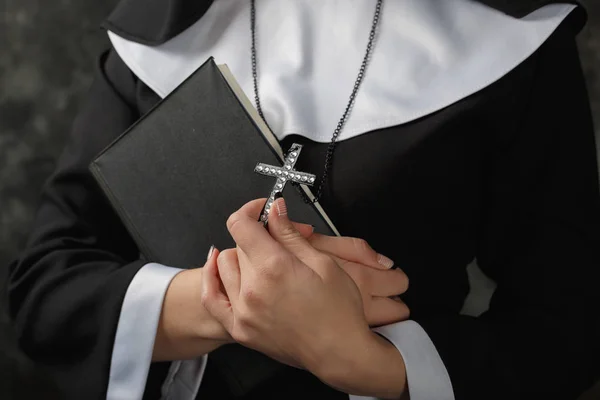 Young nun in a robe holding  bible and  cross against the dark wall. Close-up. Woman hugging  book