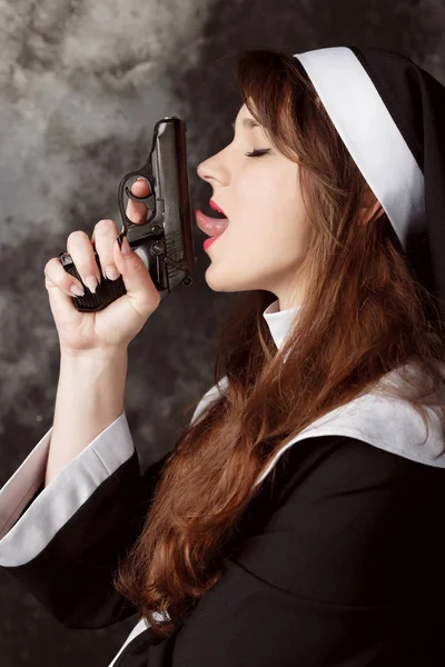 Attractive sexy nun with dark hair holding a gun and licking his tongue in a dark room