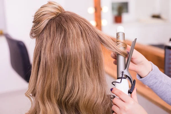 Woman hairdresser making hairstyle using curling iron for long hair of young female with smartphone in beauty salon