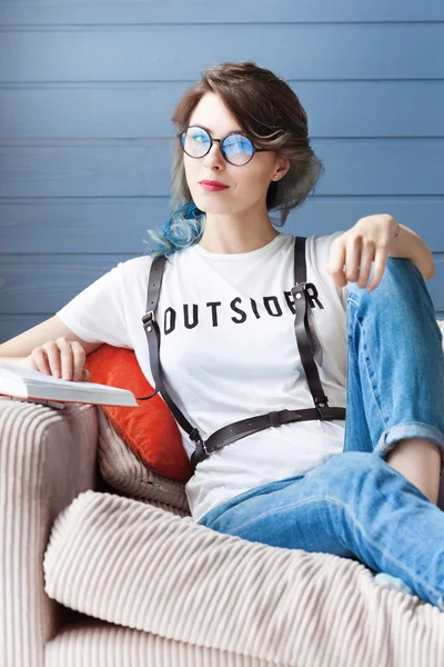 Beautiful young girl with fancy blue hair and round glasses. She is smart and happy. She is sitting on the sofa, reading orange book