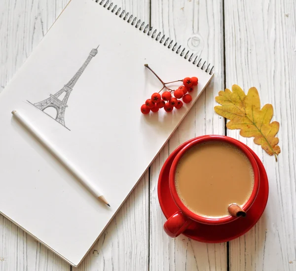 A beautiful romantic photo with a cup of coffee, a hand drawing of an Eiffel Tower in a sketchbook, an autumn leave and ash berries on a white wooden surface