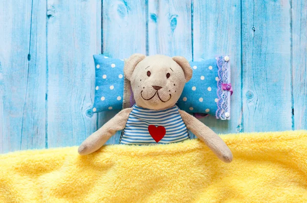 Childrens soft toy teddy bear in bed with thermometer and pills, take the temperature of a mercury glass thermometer. On a blue wooden background with a yellow blanket. Playing in hospital.
