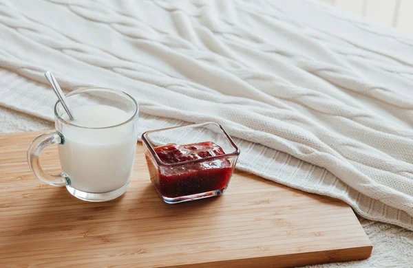 Hot milk in a glass cup and raspberry jam on a wooden board. Treatment of hot drink. Treatment of folk remedies in bed.