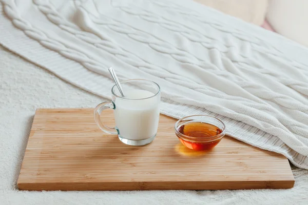 Hot milk in a glass cup and honey on a wooden board. Treatment of hot drink. Treatment of folk remedies in bed.