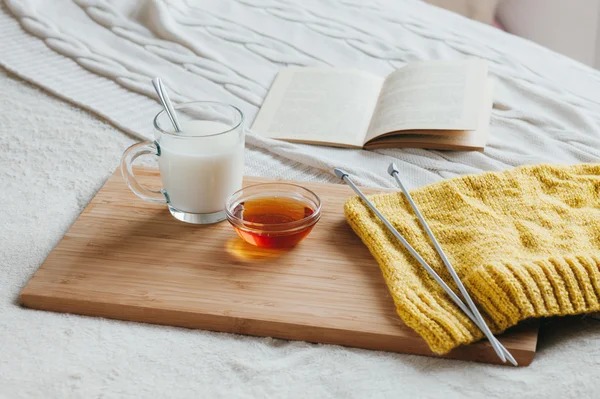 Hot milk in a glass cup and honey on a wooden board. Treatment of hot drink. Treatment of folk remedies in bed. Knitting needles and yarn for knitting. The book to read.