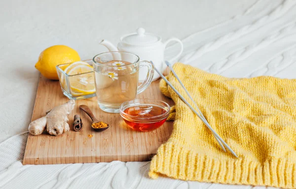 Herbal tea with chamomile flowers, turmeric and honey on a wooden board. Treatment of hot drink with ginger. Treatment of folk remedies in bed. Knitting needles and yarn for knitting.