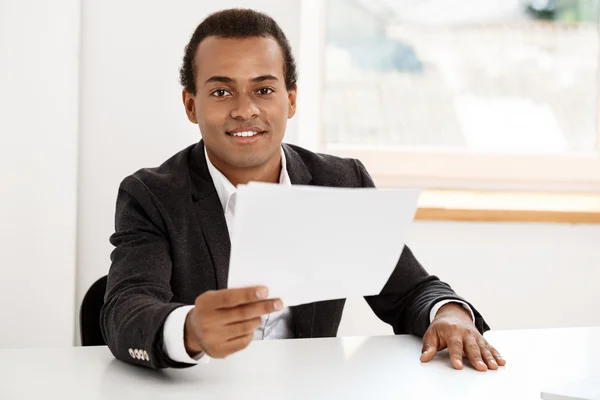 Young successful african businessman smiling, holding paper, sitting at workplace.