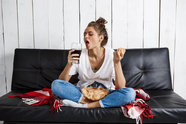 Girl eating chips, drinking soda, watching tv, sitting at sofa.