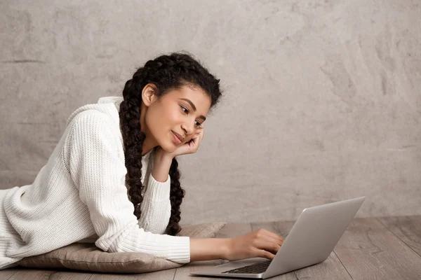 Beautiful african girl smiling, typing on laptop, lying on floor.