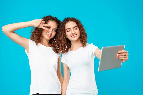 Two girls twins making selfie on tablet over blue background.