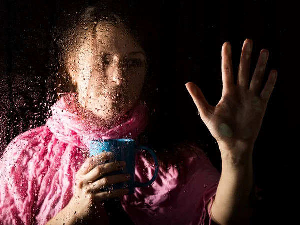 Young sad woman portrait behind the window in the rain with rain drops on it. girl holding a cup of hot drink