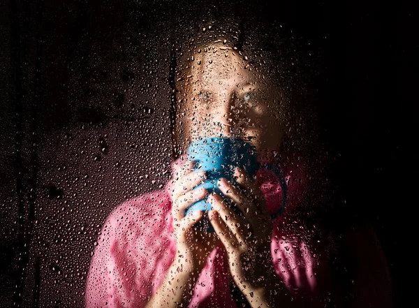 Young sad woman portrait behind the window in the rain with rain drops on it. girl holding a cup of hot drink