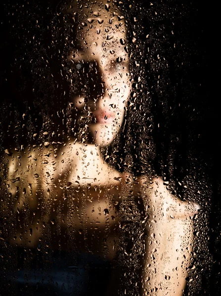 Sexy young woman, posing behind transparent glass covered by water drops. melancholy and sad female portrait