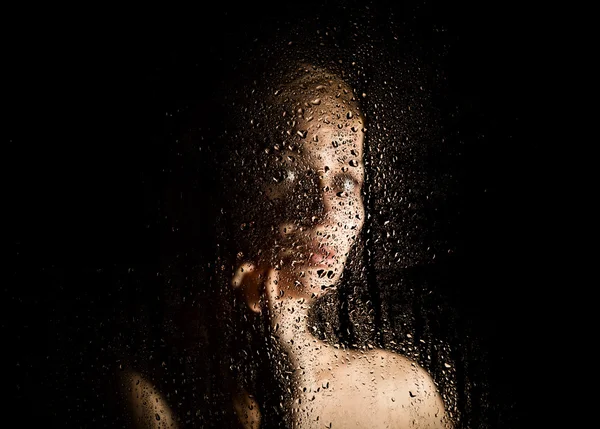Sexy young woman, posing behind transparent glass covered by water drops. melancholy and sad female portrait