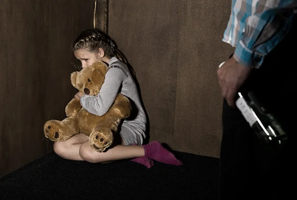 Sad little girl sitting against the wall in despair. In his hands he holds an teddy bear. drunken man threatens.