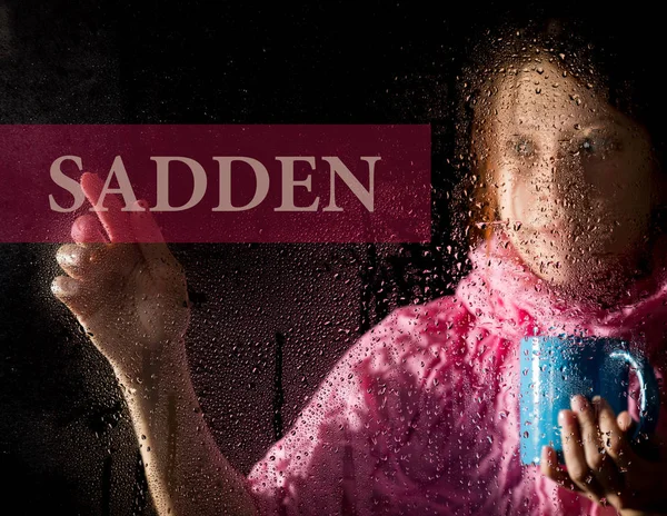 Sadden written on virtual screen. young woman melancholy and sad at the window in the rain, she holding a cup of hot coffee or tea