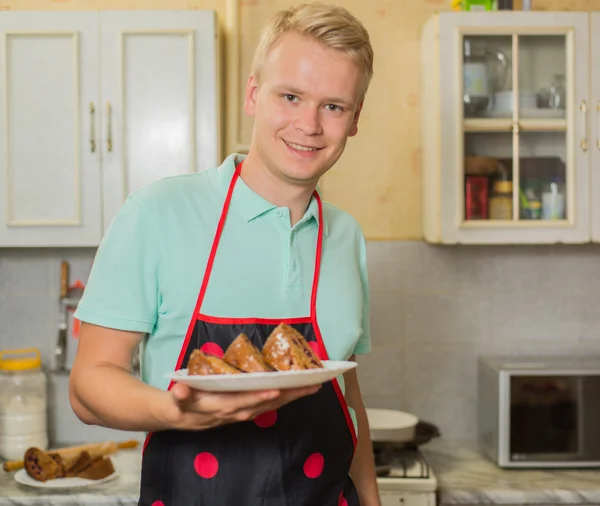 Smiling young male chef in an apron holding a cake, standing in home kitchen