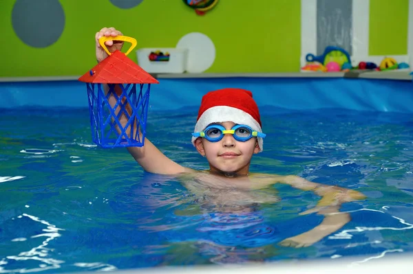 Child in hat Santa Claus swims in the pool with gift in hand on blue background and looks at me. Portrait. The view from the top. Horizontal orientation