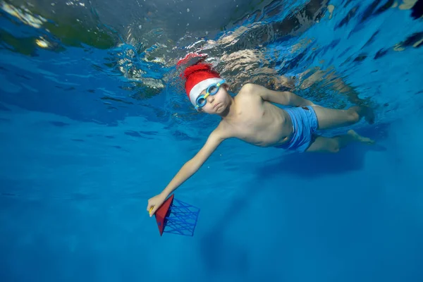 The boy in the hat of Santa Claus swims underwater with a toy in hand on blue background. The view from under the water at the bottom. Horizontal orientation