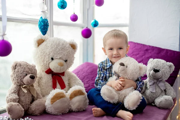 A little boy sits on a windowsill, surrounded by teddy bears. Happy boy embraces white toy bear