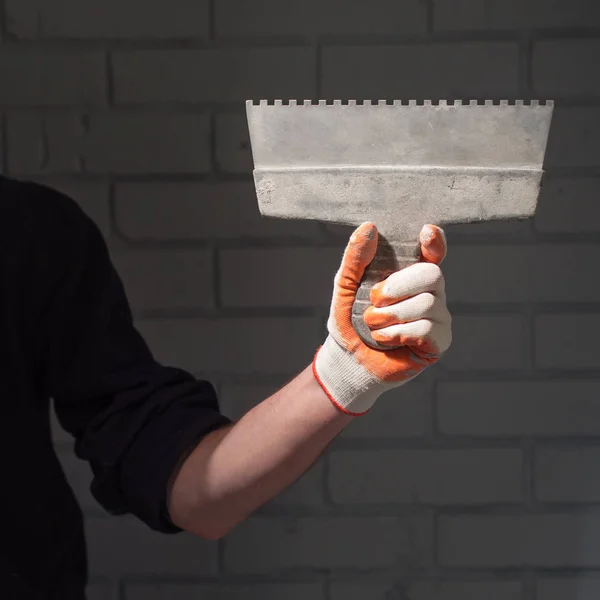 Male left arm holding large putty knife on wide abstract linear patterned textured brick wall background.