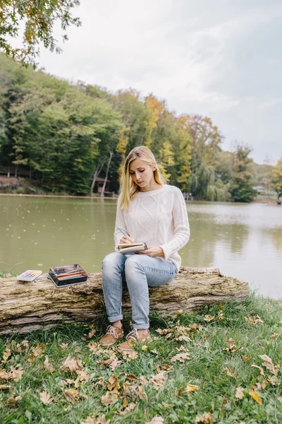 Pretty young woman in white sweater and blue jeans draws in a notebook with colored pencils sitting on trunk of a dry tree on a background of lake