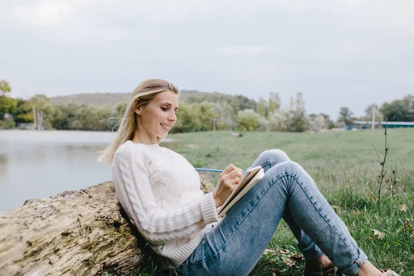 Pretty smiling young woman in white sweater and blue jeans draws in a notebook with colored pencils sitting on the grass and leaned into the trunk of a dry tree on a background of lake