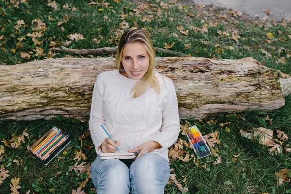 Pretty smiling young woman in white sweater and blue jeans draws in a notebook with colored pencils sitting on the grass and leaned into the trunk of a dry tree