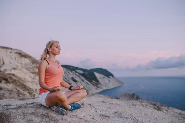 Young woman doing yoga lotus position with eyes closed outdoor on the background mountain peak cliff and sea. Sport lifestyle travel concept