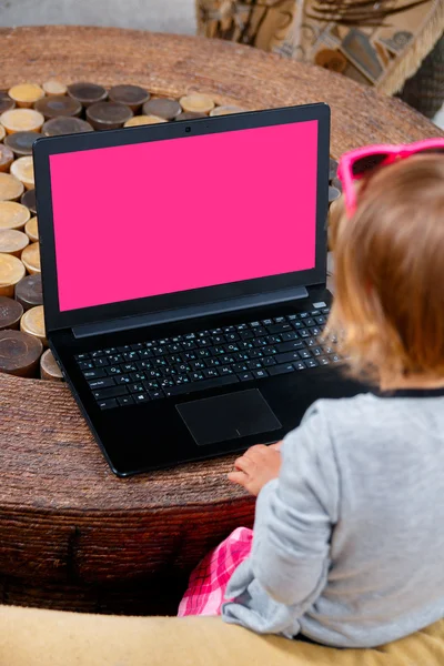 Little girl using laptop. Unrecognized child in pink skirt and  sunglasses, grey top sitting at the sofa playing, typing  on the table. Looking  the screen. back view