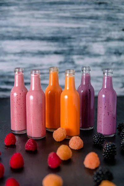 Six bottles with smoothies and raspberry, red, yellow, blackberry on black table grey background. Milk shake in glass jar berries. diet or vegan food concept, fresh vitamin.