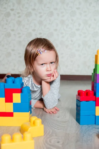 Serious Child girl lying near bright plastic construction blocks. Toddler playing on the floor. Developing toys. Early learning.