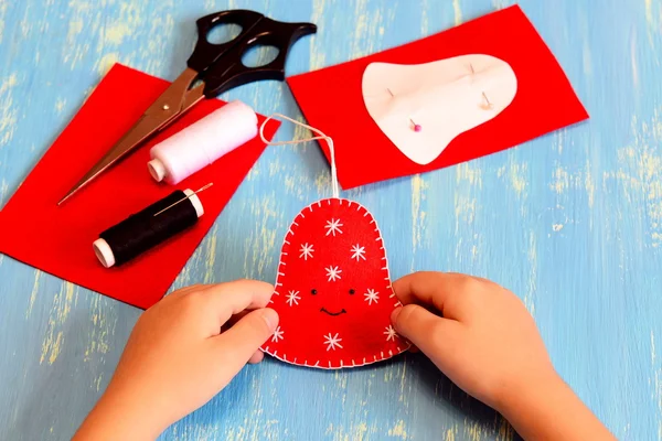 Child holds a Christmas bell toy in his hands. Child shows a felt bell. White and black threads, scissors, paper pattern, felt sheets on a wooden table. Easy Christmas art and craft idea for kids