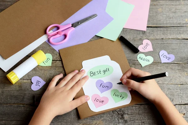 Child doing a birthday card. Child holds a black marker in hand and writes. Paper greeting card with jar, hearts and wishes. Scissors, colored paper sheets, glue stick on wooden background. Kids art