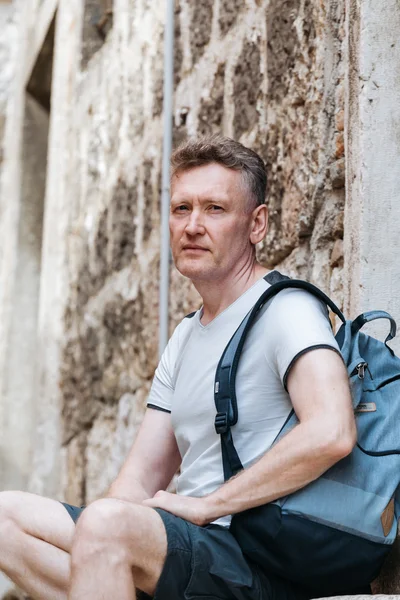 Stylish tourist. Man dressed in a white shirt and shorts with backpack over his shoulder. Sitting on the steps of European city