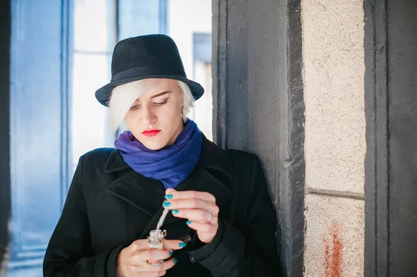 Portrait of young beautiful woman with white hair, in a black coat, a skirt and a black hat, smoking an electronic cigarette, runs vape juice electronic cigarette. He holds a mechanical mod with RDA.