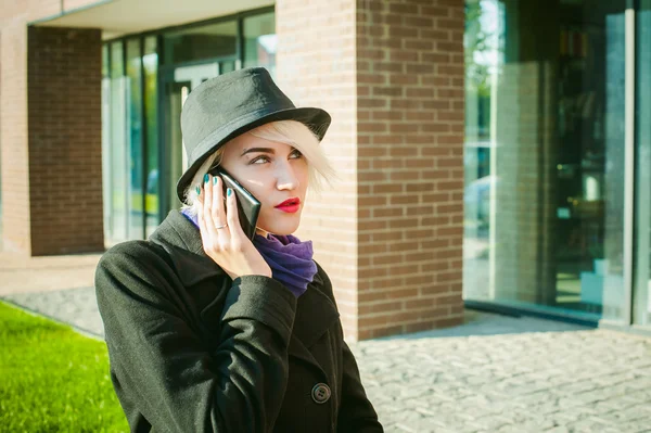 Portrait of young beautiful woman with white hair, in a black coat, a skirt and a black hat, talking on cell phone on the street. telephone communication, liaison, pleasant conversation about the business