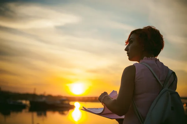 Woman with dyed red hair in a pale pink dress with white backpack, signed important documents sitting outdoors near the river at sunset