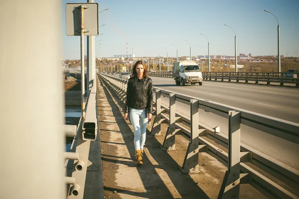 Girl goes on road bridge. lonely young woman in black Leather jacket autumn, walking over the bridge which goes transport.
