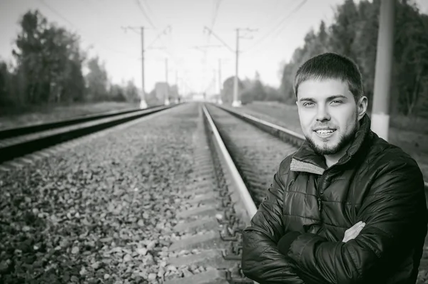 Young man sitting on tracks. bearded guy in jacket and jeans, sitting on rails and sleepers on rail until train arrived. against background of the autumn landscape and gravel, expects the new travel