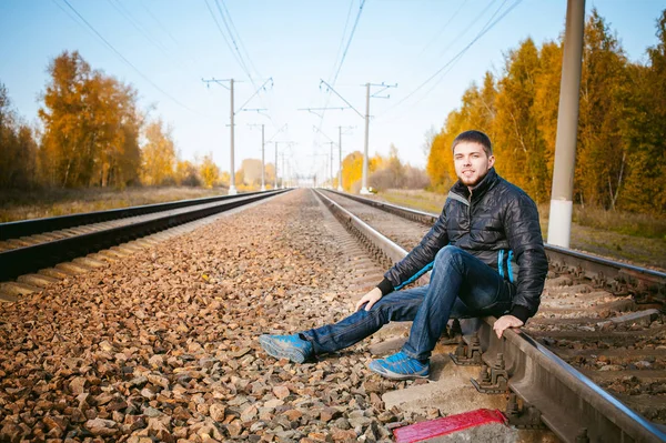 Young man sitting on tracks. bearded guy in jacket and jeans, sitting on rails and sleepers on rail until train arrived. against background of the autumn landscape and gravel, expects the new travel