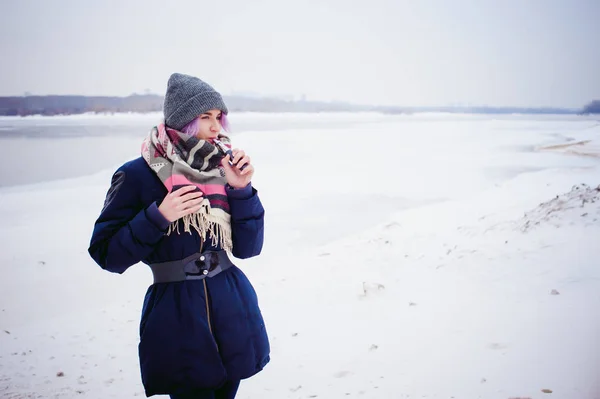 Vaping girl. winter street portrait of a woman hipster, purple-dyed hair, a gray knitted hat and scarf. woman smokes an electronic cigarette in the street near the snow-covered river