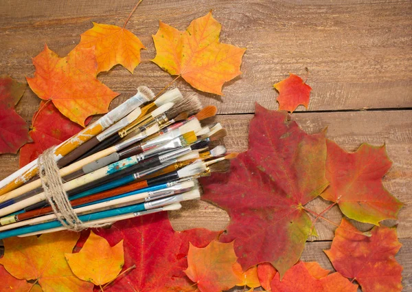 Paints, brushes, autumn leaves on wooden background.
