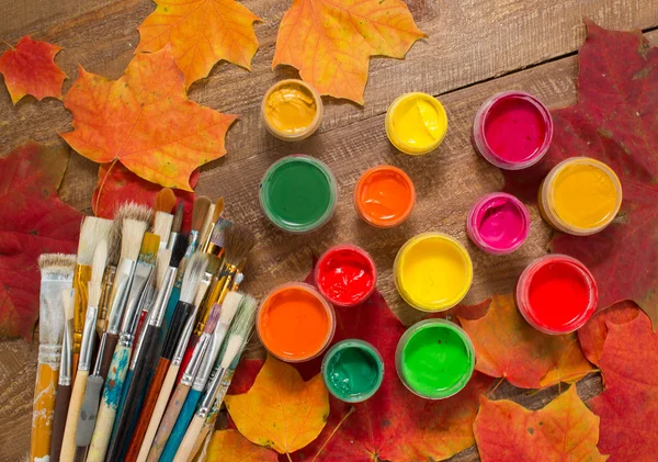 Paints, brushes, autumn leaves on wooden background.