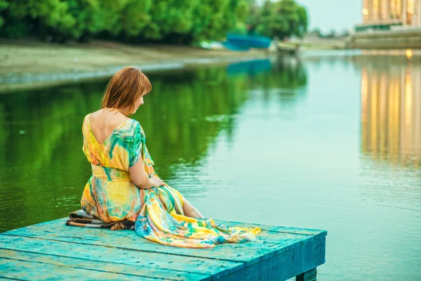 Red-headed woman in bright summer dress with open back sitting on the wooden pier at the river bank in the city