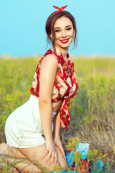Beautiful young girl with perfect tempting smile wearing red blouse, white shorts and headband sitting in the field