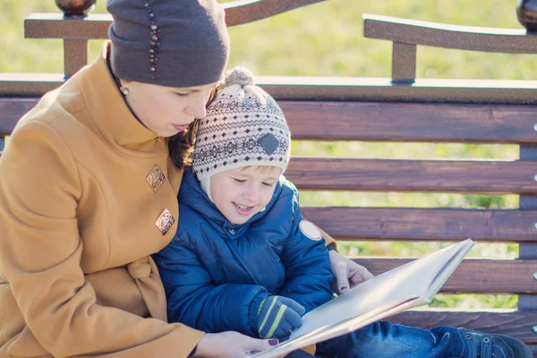 Young woman and little boy sitting on the bench in autumn Park and reading book. Family enjoying time together. Mother teaches son