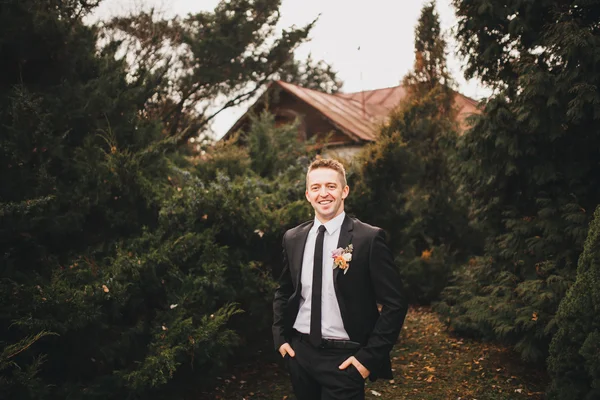 Beautiful stylish young groom in black wedding suit white jacket shirt tie, standing outdoors in the woods, on a natural background